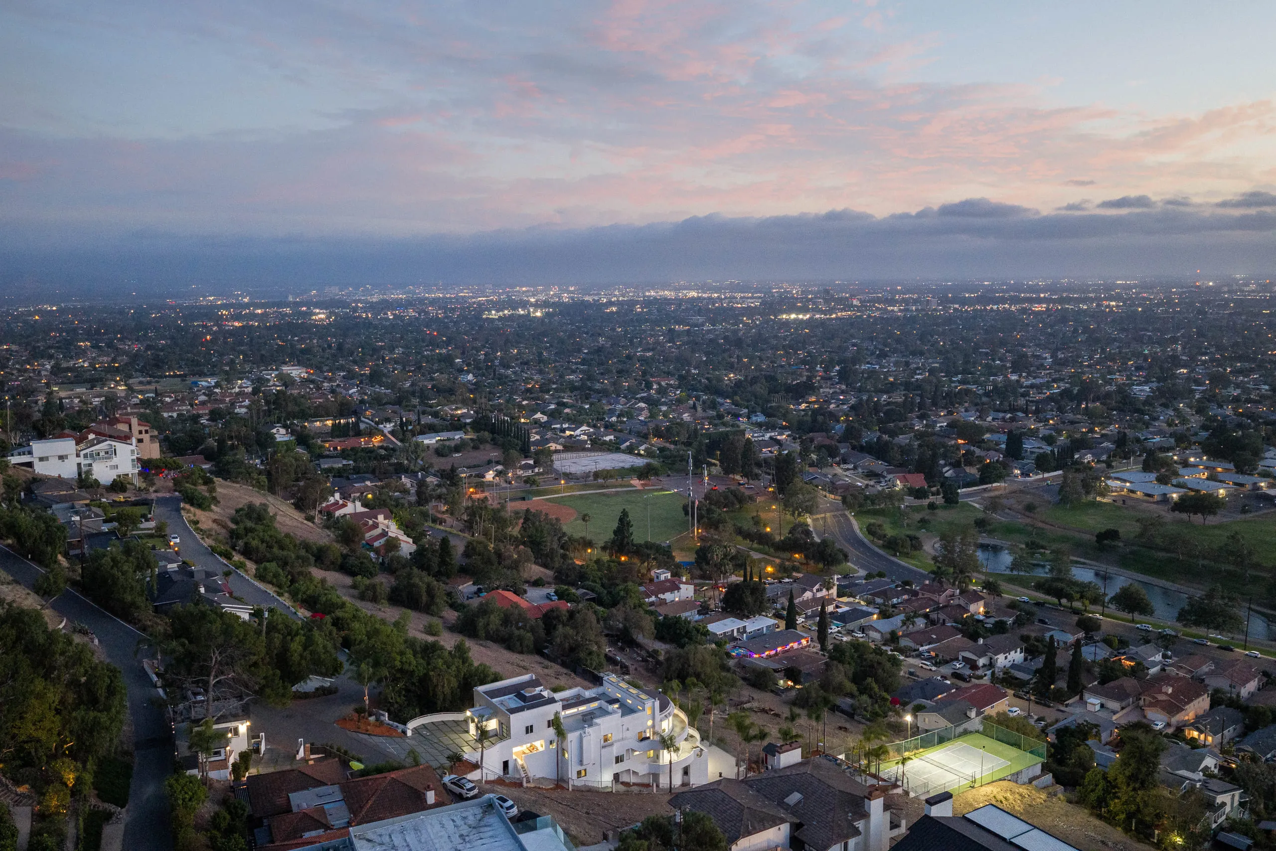 City lights across the basin at dusk, the lighted court glowing below the house